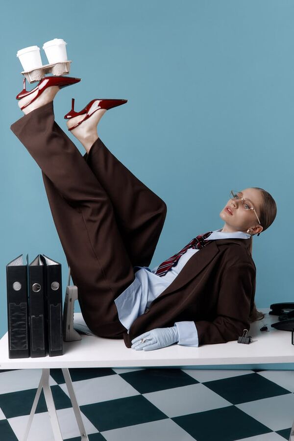 Model in a brown suit lying on a desk balancing coffee cups on red heels