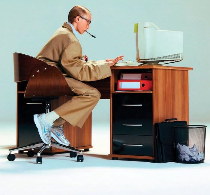 Man in a camel suit with a pen in his mouth typing at a retro office desk